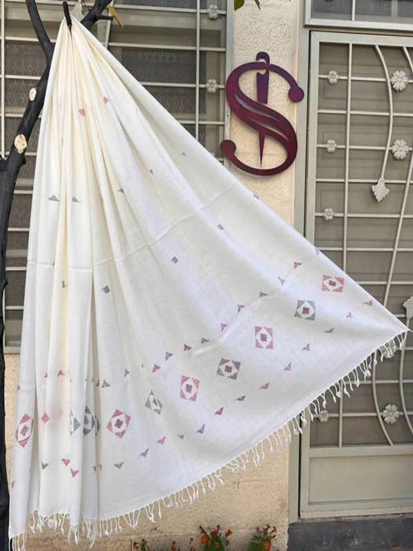 A white handmade woolen shawl made in swat kpk pakistan by hand operated looms or handlooms with hand embroidery done at the time of waving   in red, green, color, draped over a black tree branch in front of a ware house  with a metal gate and a dark purple saidaar handmade product logo sign.